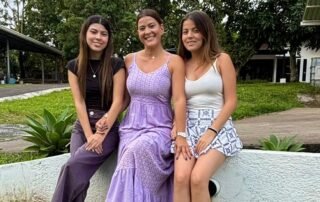 Retrato panorámico de Fabi Herra y sus dos hijas, Sofi y Lucía, sonriendo juntas en un banco de madera en un jardín al aire libre.